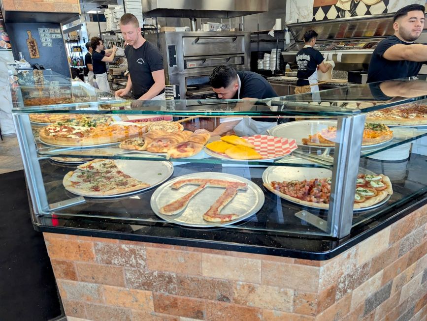Glass pizza display case at Anthony Francos in Springfield, New Jersey, featuring various pizza slices and baked goods including a slice shaped like the Pi (π) symbol. Staff members are actively working behind the counter, with ovens and topping stations visible in the background.