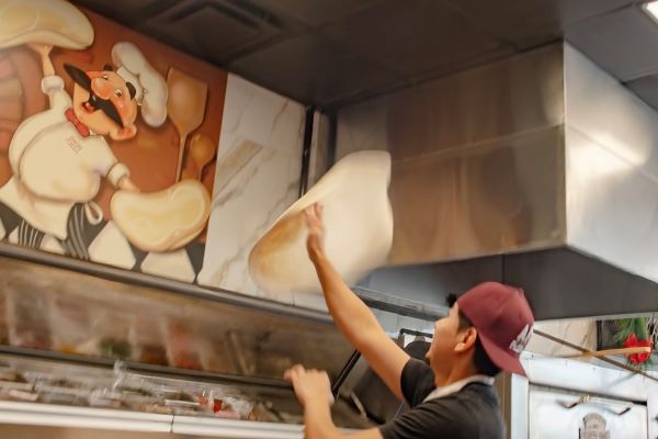 A man in a black shirt and hat is tossing a pizza high in the air at Anthony Francos in Springfield, NJ.